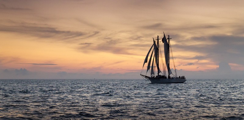 A sailing ship with tall masts and sails is silhouetted against a colorful sunset sky over calm ocean waters.