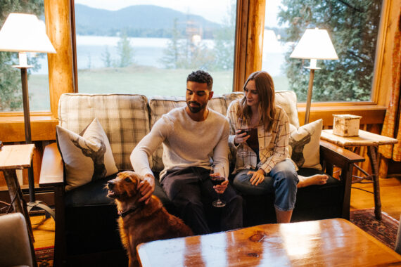 A couple sits on a sofa in a cozy room at Lake Placid Lodge, each holding a glass of wine. The man gently pets a dog beside him as large windows reveal a scenic view of the lake and mountains. Soft lighting enhances the warm, inviting atmosphere.