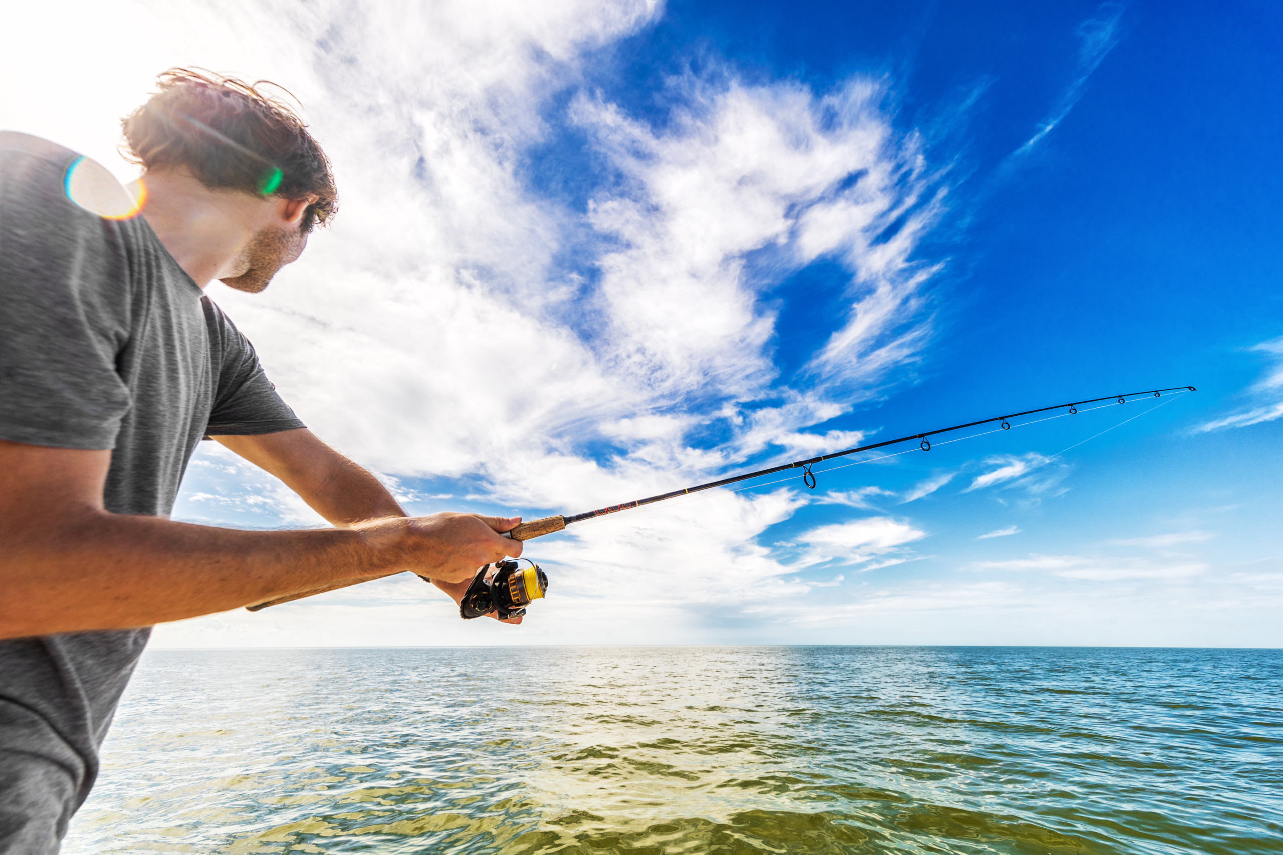 A person wearing a gray t-shirt is enjoying Hutchinson Island fishing in a vast body of water under a bright blue sky with scattered clouds. They are holding a fishing rod and facing the horizon where the water meets the sky. Sunlight creates a lens flare on the left side.