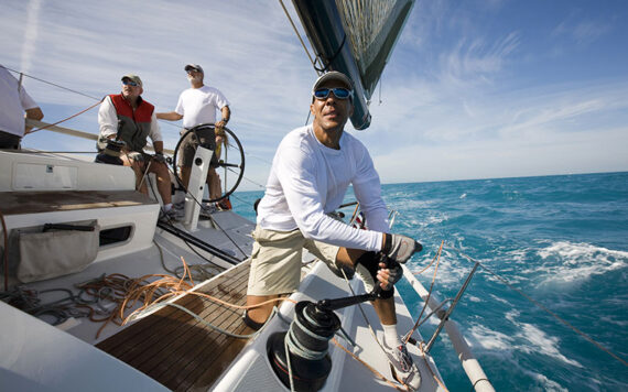A group of people sailing on a boat in the ocean under a clear blue sky. One person is adjusting a sail while others are navigating and observing. The water is a vibrant blue, and the boat is angled slightly amidst small waves.