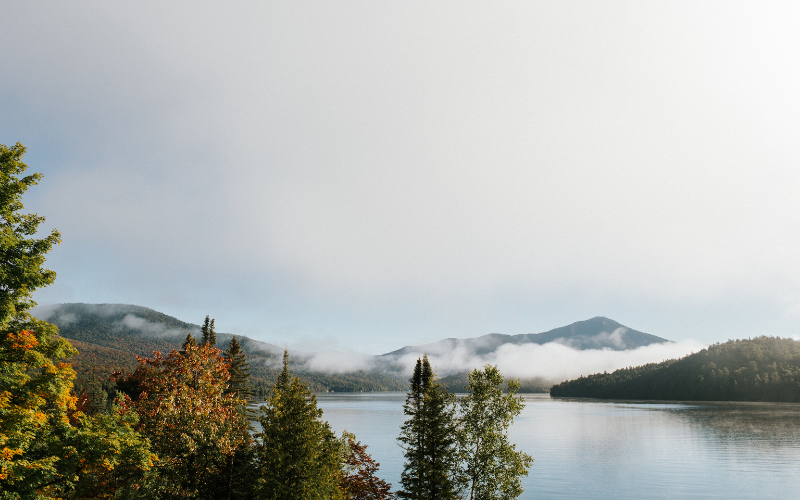 A serene lake, reminiscent of Lake Placid, is surrounded by forested hills under a clear sky. Mist floats above the water, and the trees display a mix of green and autumnal colors, creating a peaceful natural landscape that hints at things to do in this picturesque setting.
