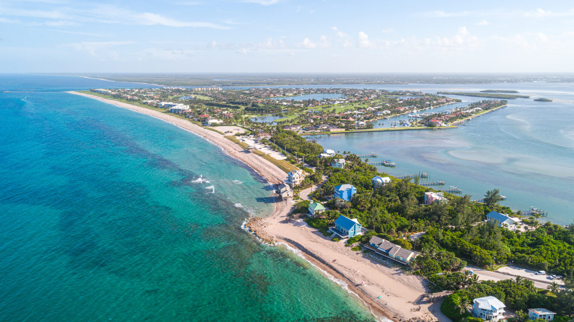 Aerial view of a coastal landscape featuring a sandy beach bordered by clear turquoise waters on the left and a residential area with greenery and waterways to the right, under a clear blue sky.