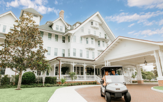 A couple drives a golf cart past a large, elegant white Victorian-style building with multiple gables and a covered driveway. Its a sunny day with a few clouds in the sky, and the surrounding landscaping is lush and well-maintained.
