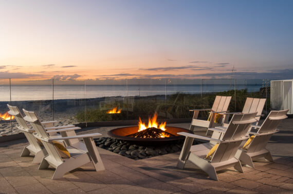 Outdoor seating area with white chairs surrounding a fire pit, set against a beach backdrop at sunset. The sky is colorful, and a glass barrier separates the area from the beach.