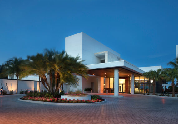 Modern building with a clean, white facade and large glass entrance, surrounded by well-maintained palm trees and a flower bed. The sky is clear, creating a peaceful evening ambiance.