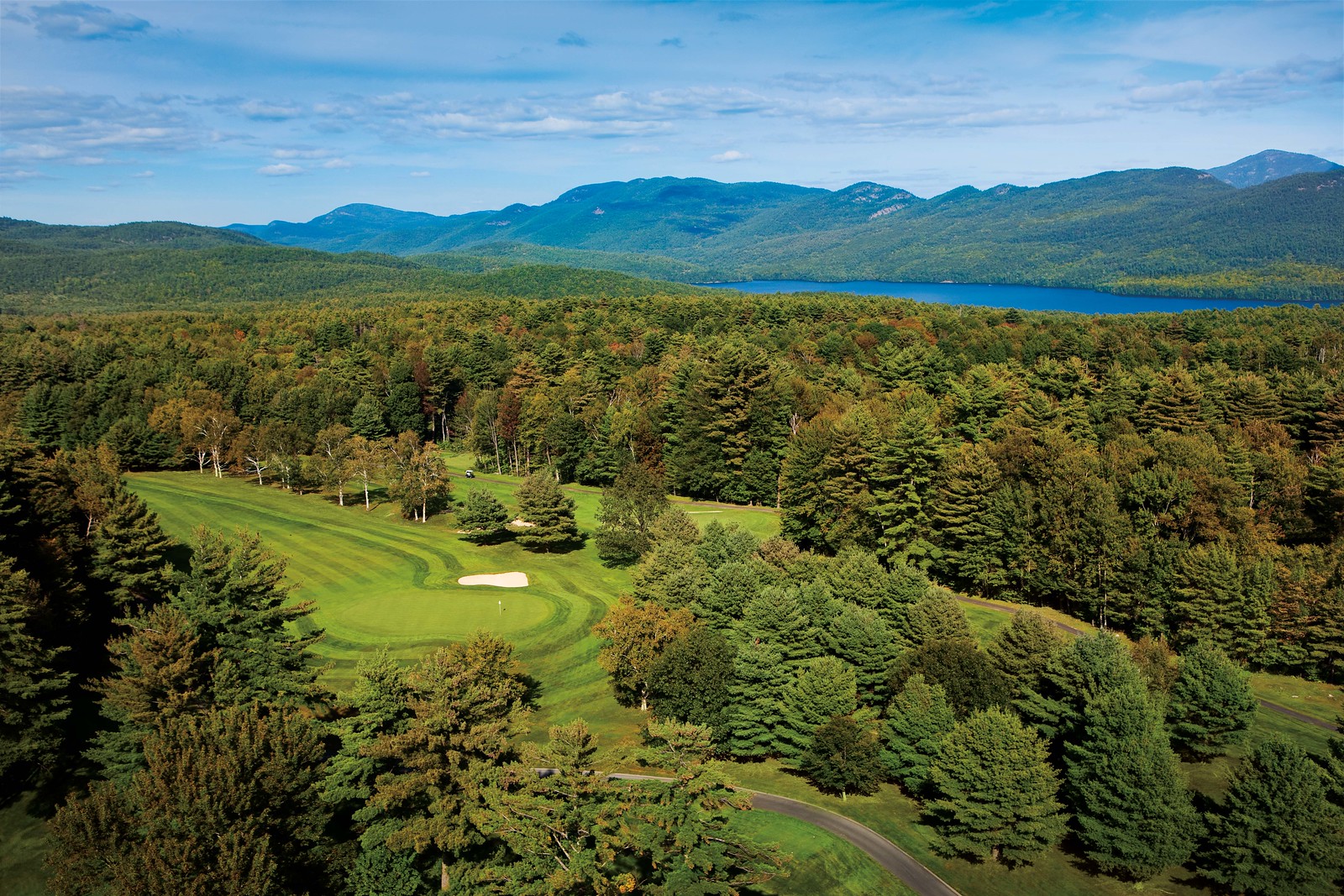 A picturesque aerial view of a lush green golf course surrounded by dense forests and rolling hills. In the distance, a serene blue lake can be seen beneath a clear, blue sky with mountains on the horizon.