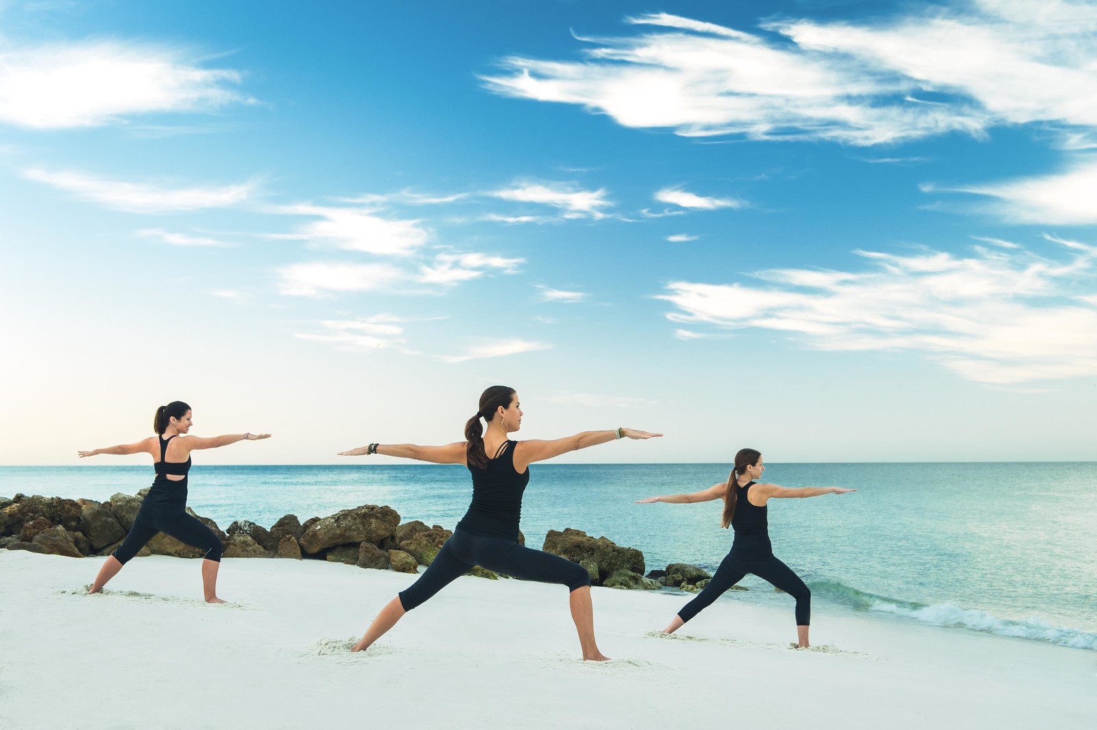 Three people practice yoga in a warrior pose on a sandy beach, with a calm ocean and a blue sky with scattered clouds in the background.