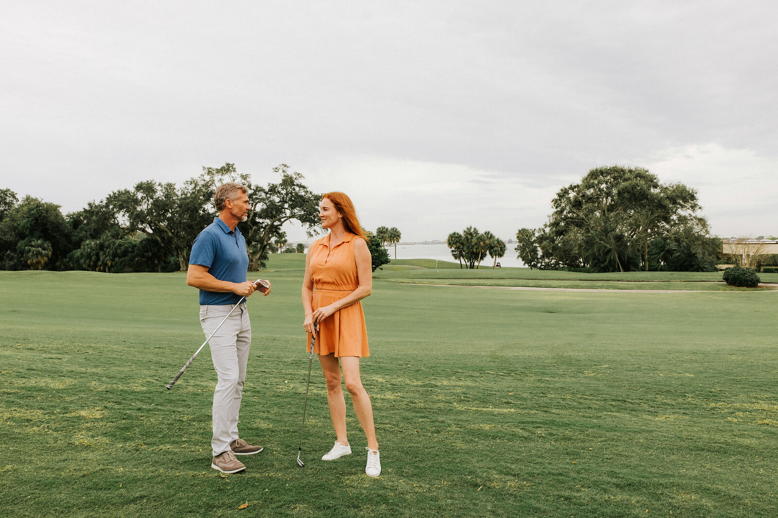 A man and woman are standing on a golf course, engaged in conversation. The woman is wearing an orange dress and holding a golf club, while the man is in a blue shirt and light pants. In the background are trees and a distant body of water.