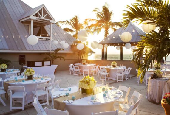 Outdoor rooftop event space at sunset with round tables and white chairs. Tables are decorated with white flowers and small turquoise boxes. Palm trees and string lights with white lanterns enhance the tropical ambiance.