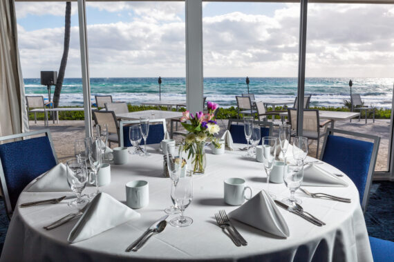 A round table set for a formal meal, with white tablecloth, folded napkins, glassware, and a floral centerpiece. The setting overlooks a scenic ocean view through large windows, with outdoor seating visible on a patio.