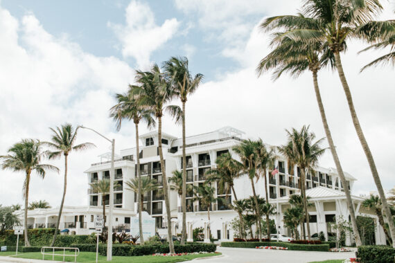A modern white hotel is surrounded by tall palm trees under a partly cloudy sky. The building has multiple balconies and a circular driveway with landscaped greenery.