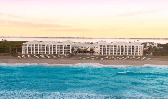 Aerial view of a large beachfront hotel at sunset. The hotel is surrounded by palm trees, and rows of white cabanas line the sandy beach. The calm ocean waves gently lap the shore, while the sky glows with warm hues.