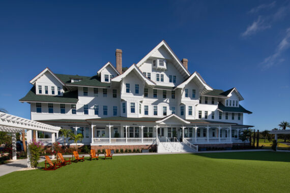 A large, white, multi-story building with gabled roofs and numerous windows stands under a clear blue sky. It has a wide porch and a well-manicured green lawn in front, with several wooden chairs and a pergola on the side.