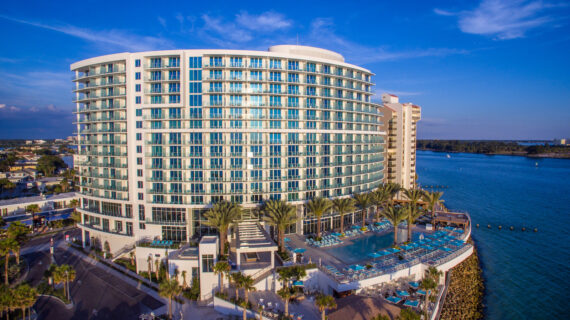 An aerial view of a large, modern beachfront hotel with curved architecture and glass balconies. The hotel overlooks a calm blue ocean, with a pool area surrounded by palm trees and sun loungers on a sunny day.