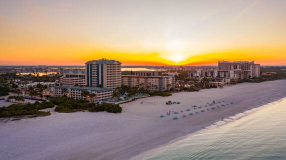 A picturesque sunset over a coastal city with sandy beaches. High-rise buildings line the shore, and the sun sets on the horizon, casting a warm glow over the ocean and sky. Rows of lounge chairs are set up on the beach.