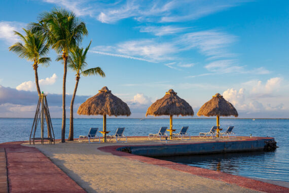 A tranquil beach scene with an empty pier extending into the ocean. Three thatched umbrellas shade lounge chairs. Two palm trees stand to the left under a vibrant blue sky with scattered clouds.