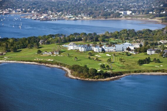 Aerial view of a scenic coastal area featuring a golf course with lush green fairways and sand bunkers. Buildings are nestled among trees, and a calm body of water surrounds the landscape, with a distant city and boats visible on the horizon.