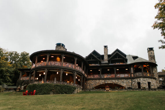 A large, rustic stone mansion with multiple balconies and lit windows stands on a grassy slope. The building features round and angular sections with wooden railings, surrounded by trees under a cloudy sky.