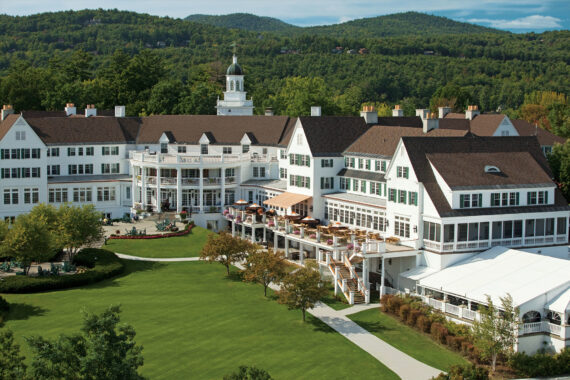 A grand white resort with a sprawling lawn, surrounded by lush greenery and mountains. The building features multiple levels with balconies and a central tower. Outdoor seating areas are visible, and the sky is clear and sunny.