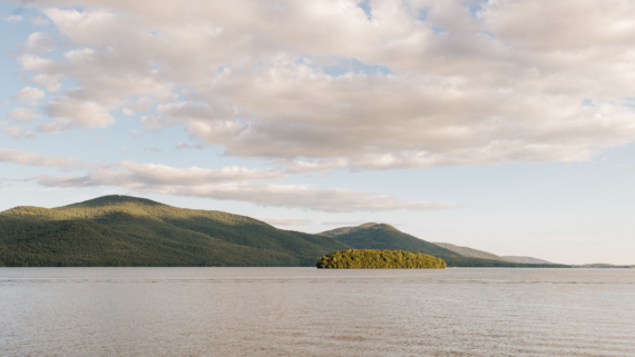 A serene landscape featuring a calm lake with a small, forested island in the center. Surrounding the lake are gentle, green mountains under a partly cloudy sky, bathed in warm sunlight.