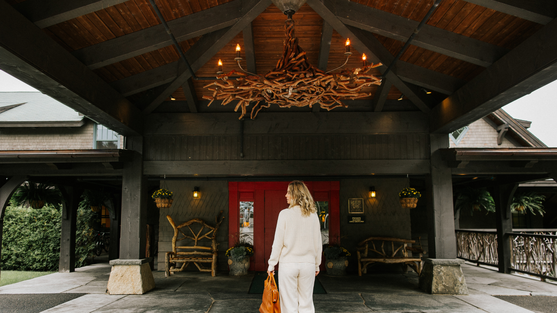A person with blonde hair and a white outfit stands under a wooden canopy, facing a building with a red door. Rustic benches and a large branch chandelier decorate the space.