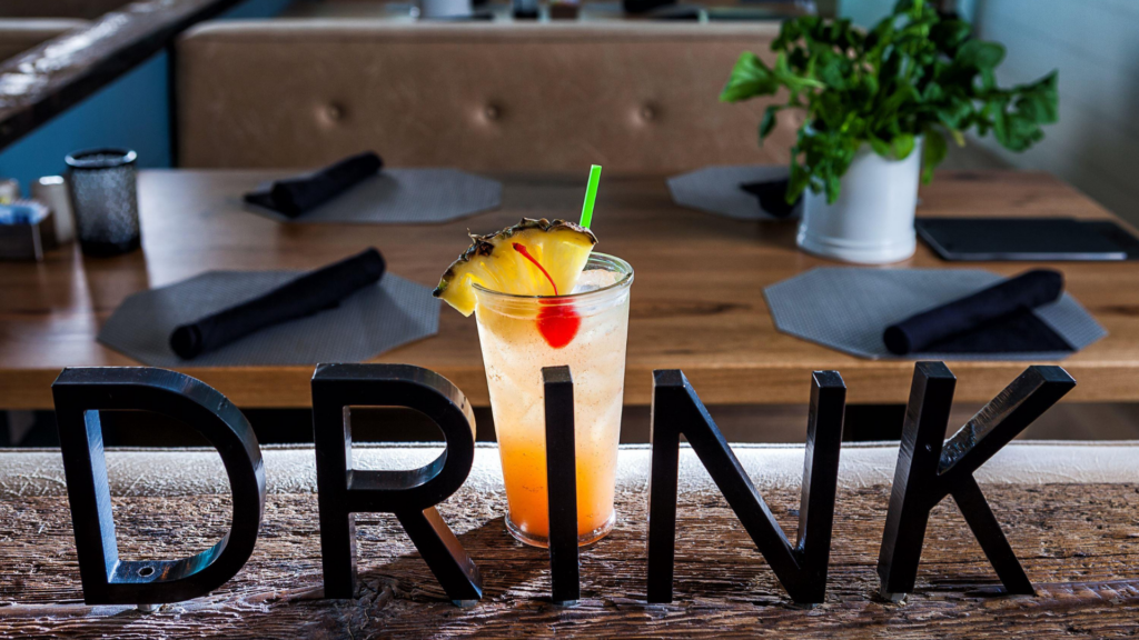 A tropical cocktail garnished with a cherry and pineapple slice sits on a rustic wooden table. In front, large black letters spell DRINK. The background features a dining table with placemats, black napkins, and a potted plant.