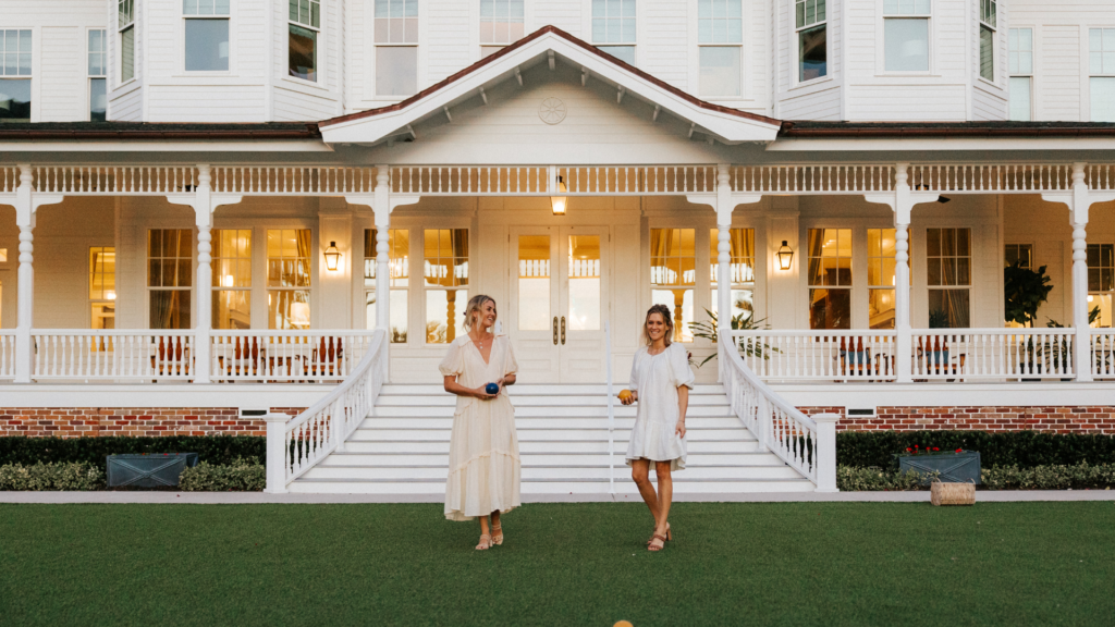 Two women in light dresses stand on a lawn in front of a large, elegant building with a wide staircase and white wooden details. They appear to be holding bocce balls, suggesting they are playing or about to play the game.