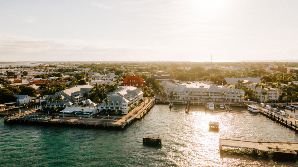 Aerial view of a coastal town at sunset, featuring a marina with boats docked along the piers. Sunlight reflects off the water, and buildings with red and white roofs are surrounded by lush greenery. The sky is clear with a warm glow.