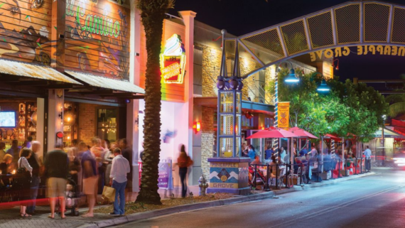 A vibrant street scene at night in a lively district with colorful storefronts and outdoor seating. People walk along the sidewalk, enjoying the bustling atmosphere. String lights and a decorative sign above the street add to the festive ambiance.