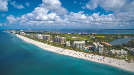 Aerial view of a coastal area with a white sandy beach lined with buildings and umbrellas. The turquoise ocean meets the shoreline under a partly cloudy sky. Residential buildings are surrounded by green landscape.