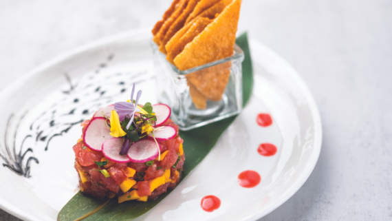 A gourmet dish featuring tuna tartare garnished with sliced radishes and edible flowers, served on a leaf with a glass holding crisp triangle chips and dots of red sauce on a white plate.