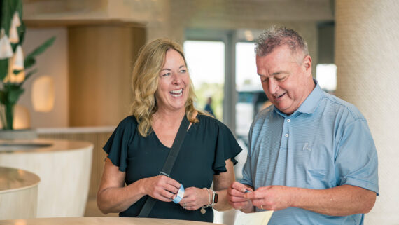 A smiling woman and man stand indoors at a counter. The woman holds a card and looks at the man, who is focused on papers in his hands. The setting appears modern and warmly lit.