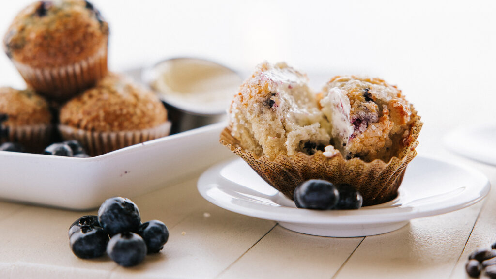 A blueberry muffin split in half with melting butter sits on a white plate, surrounded by fresh blueberries. More muffins and blueberries are in the background on a white platter.