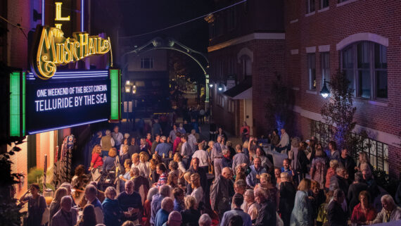 A lively crowd gathers outside The Music Hall at night, illuminated by neon signs and streetlights. The marquee reads One Weekend of the Best Cinema: Telluride by the Sea. The brick buildings and festive atmosphere suggest a special event.