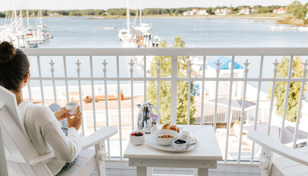 A person sits on a balcony overlooking a marina, holding a mug. A table nearby holds breakfast items, including a bowl of cereal, croissants, berries, and a French press. Boats and waterfront houses are visible in the background.