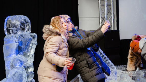 Two woman pose for a selfie in front of an ice sculpture.