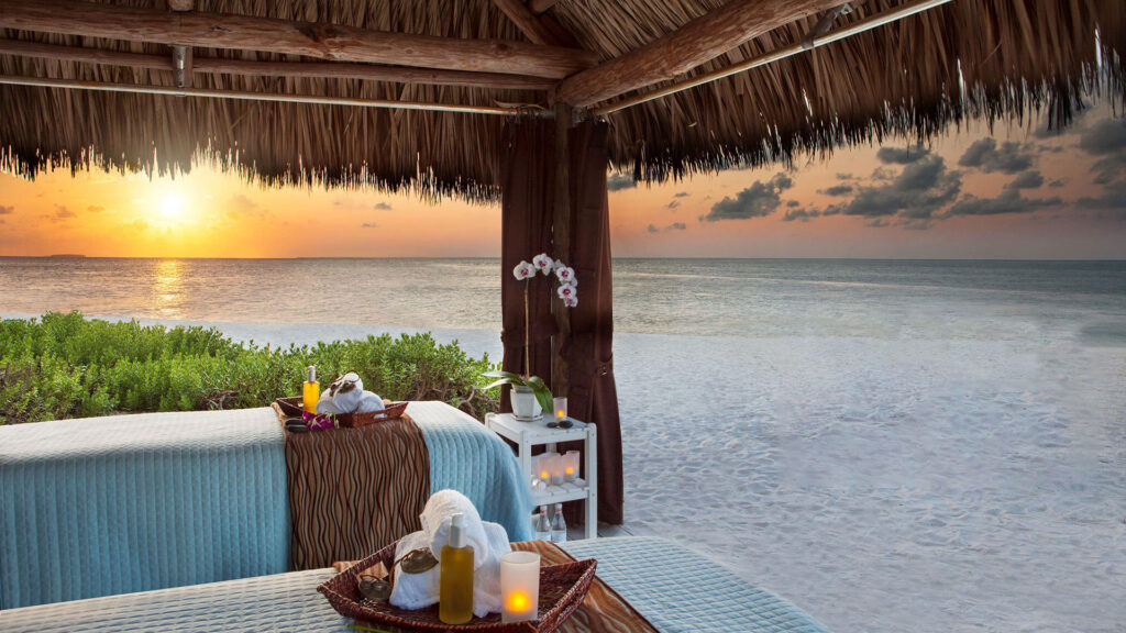Beachside hut with massage tables facing the ocean at sunset. A table holds candles and towels. The sky is orange and purple, reflecting on the water. Relaxing, serene atmosphere with a peaceful beach view.
