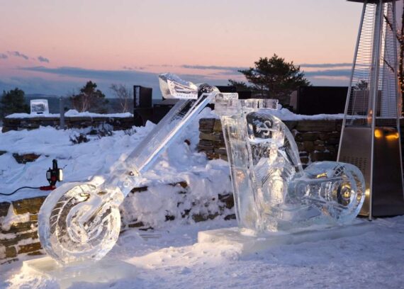 A detailed ice sculpture of a motorcycle is displayed outdoors on a snowy landscape. The sky is painted in soft hues of pink and orange from the sunset. A heater stands nearby, and trees are visible in the background.