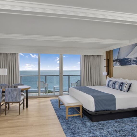 Hotel room with a large bed, blue and white decor, and a sailboat mural above the headboard. A sitting area with a table and chairs is on the left. Glass doors open to a balcony with ocean views under a clear sky.