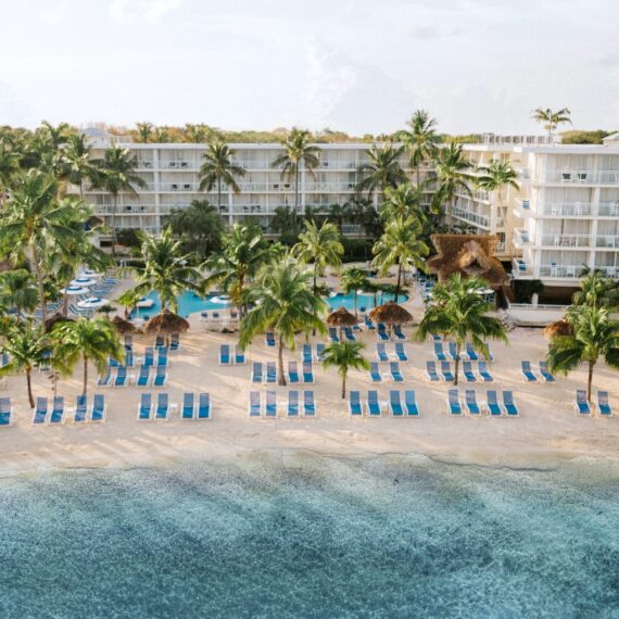 Aerial view of a beachfront resort featuring a sandy beach lined with blue lounge chairs and straw umbrellas. Behind the beach, there is a large swimming pool surrounded by palm trees and a multi-story hotel building. The sea is calm and clear.