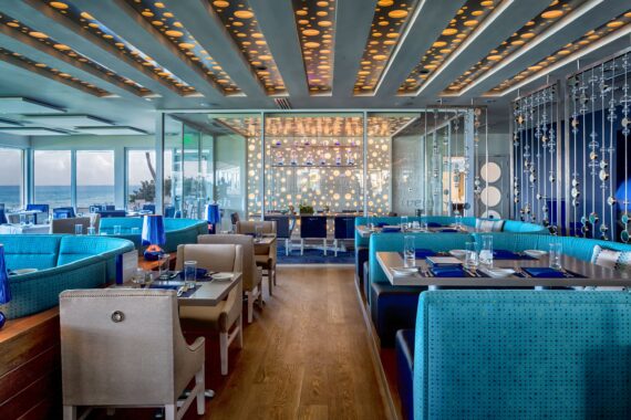 A modern restaurant interior with blue and beige seating. The ceiling features round illuminated panels, and the glass wall is decorated with circular patterns. Large windows offer a view of the ocean, enhancing the bright and airy atmosphere.
