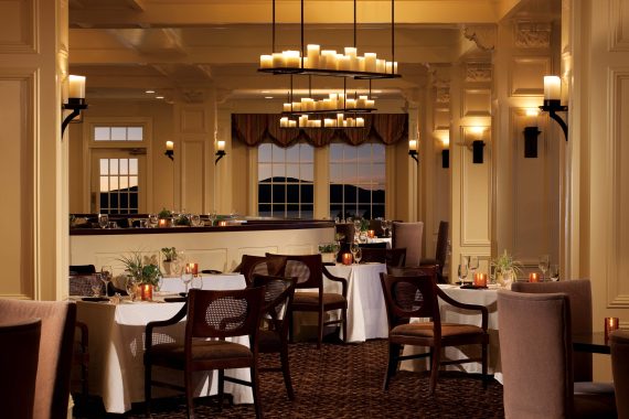 A warmly lit, elegant restaurant interior with neatly arranged tables covered in white cloths and cushioned chairs. Candles and small plants decorate the tables, and large windows reveal a scenic evening view.