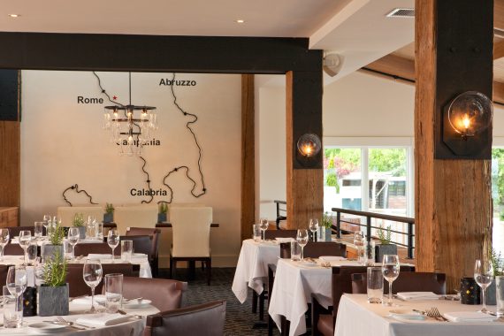 Elegant restaurant interior with white tablecloths and brown chairs. A wall displays a minimalist map of Italy featuring Rome, Abruzzo, Calabria, and Campania. A chandelier hangs from the ceiling, and large windows offer natural light.