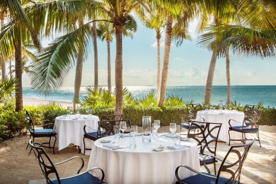 Outdoor dining area with round tables covered in white tablecloths, set with glassware and cutlery. The tables are surrounded by palm trees with a view of the ocean and blue sky in the background.