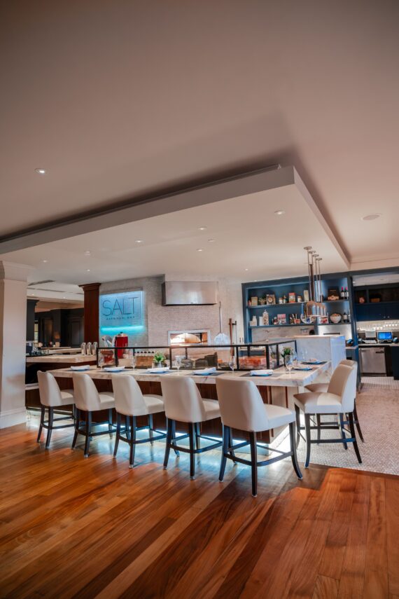 Modern kitchen space with a long dining counter lined with white leather chairs. The room features warm wooden flooring, sleek countertops, and decorative shelving. A sign with SALT is illuminated on the wall near the cooking area.