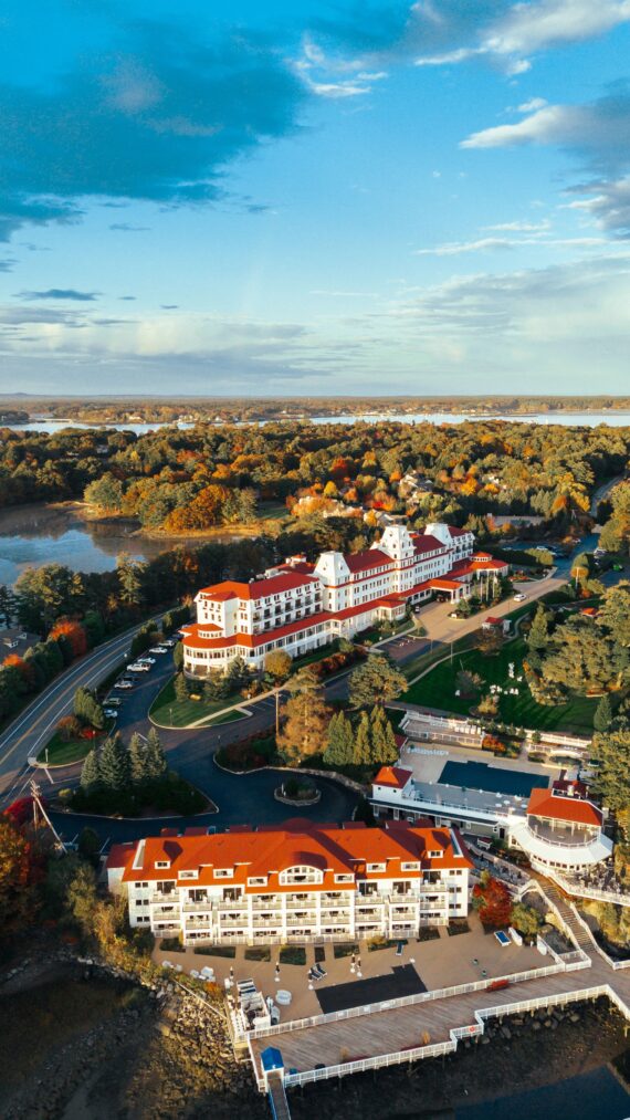Aerial view of a large, red-roofed building surrounded by lush greenery and autumn-colored trees near a body of water. The complex features several structures and a dock, creating a scenic landscape under a partly cloudy sky.