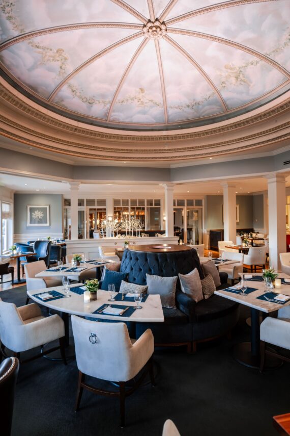 Elegant restaurant interior with a large, ornate dome ceiling painted with a sky motif. The room features plush seating around white tables, soft lighting, and decorative floral arrangements, creating a sophisticated and inviting atmosphere.