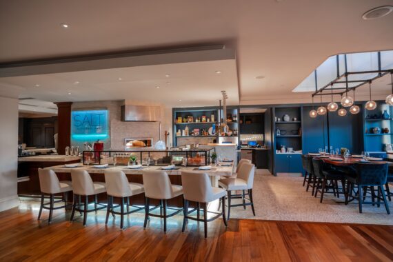 Chic restaurant interior featuring a long dining counter with white chairs, open kitchen with wood-fired oven, shelves with decor, and a cozy seating area with a round table and modern lighting fixtures. The space has a warm, inviting ambiance.