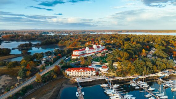 Aerial view of a coastal area with a resort featuring red roofs surrounded by trees in autumn colors. There are several boats docked at a marina, a winding road, and water bodies reflecting the sky.