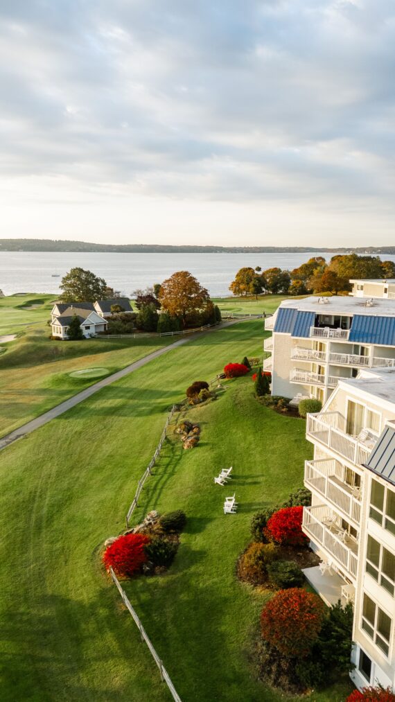 A scenic view of a lakeside resort with white buildings and blue roofs. Lush green lawns stretch to the lake, bordered by vibrant red shrubs. Wooden chairs are placed on the grass, and a clear sky enhances the tranquil ambiance.